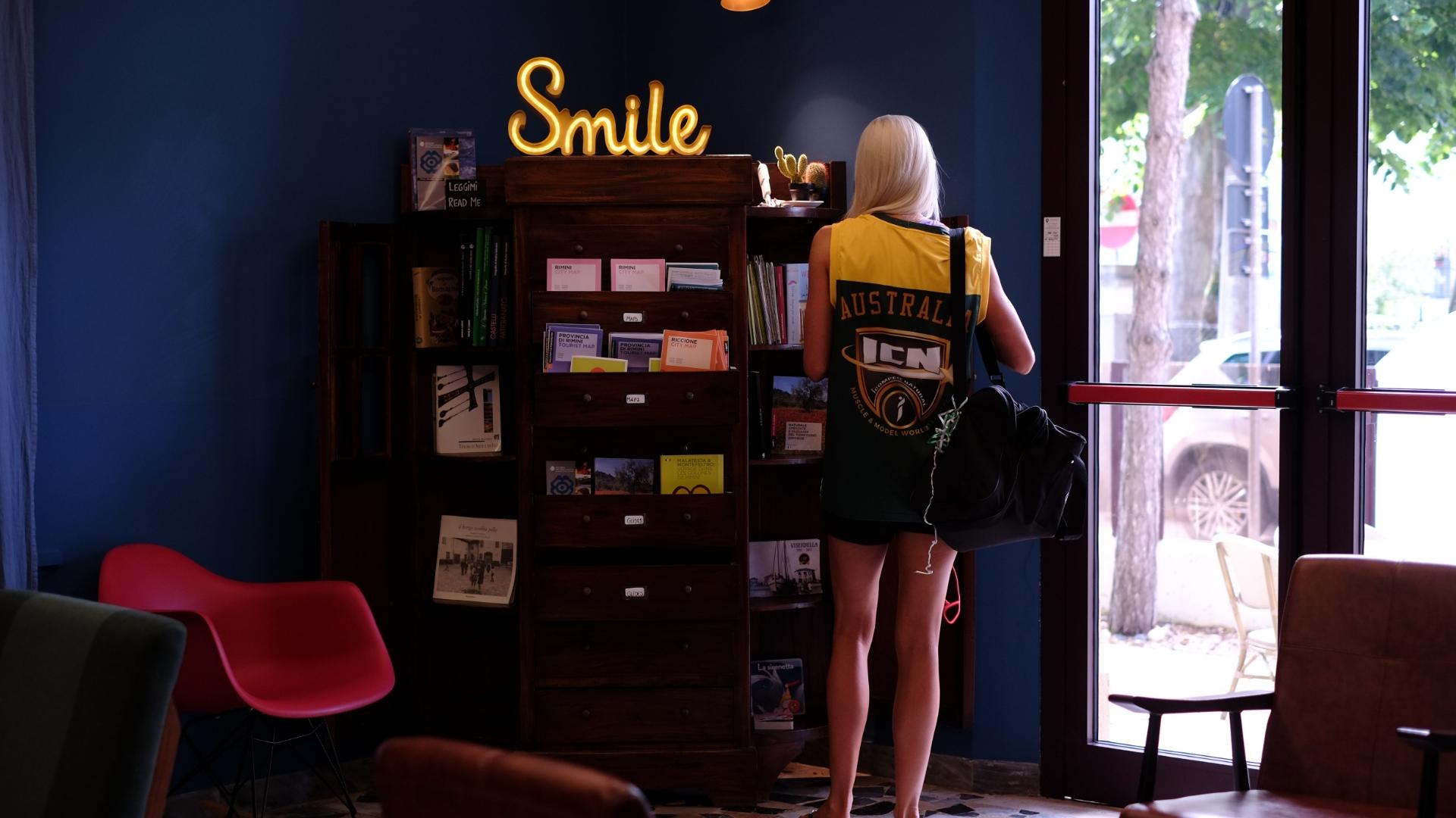 Persona in un caffè con una maglia australiana, vicino a una libreria e 'Smile'.
