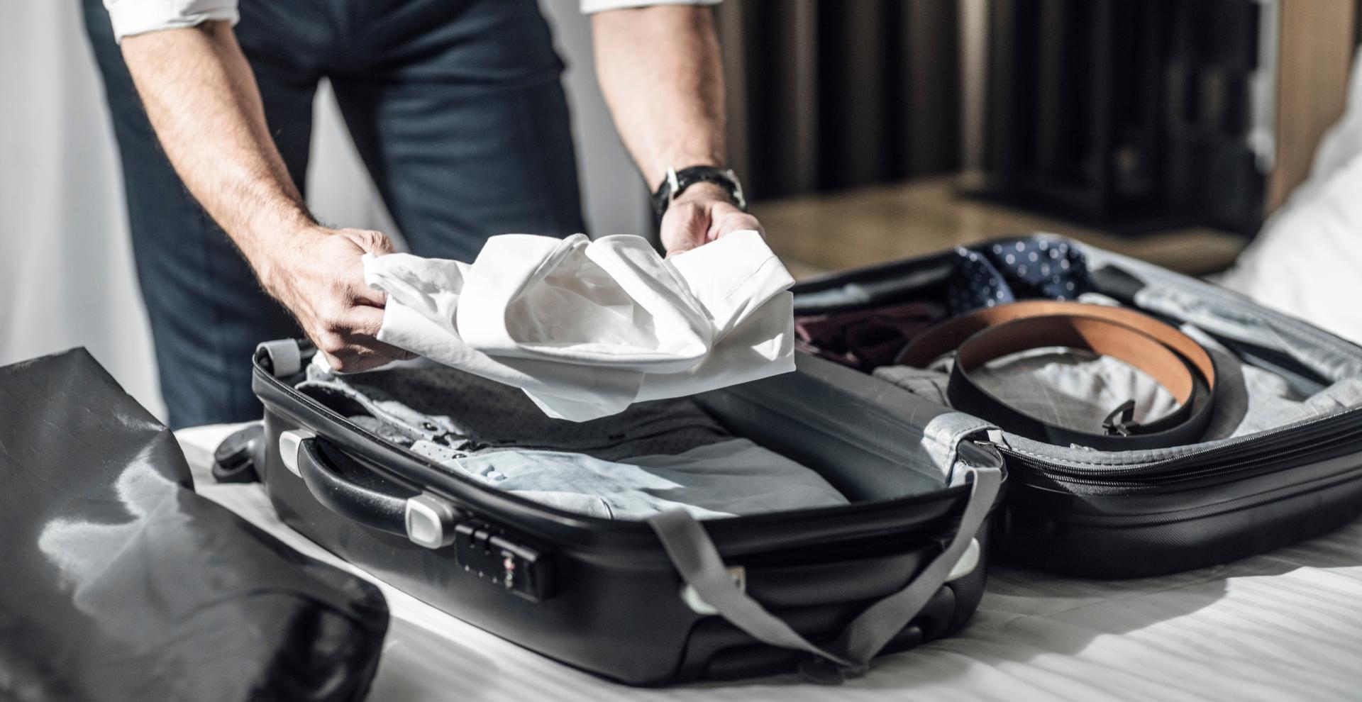 Persona che prepara una valigia con vestiti e accessori su un letto.