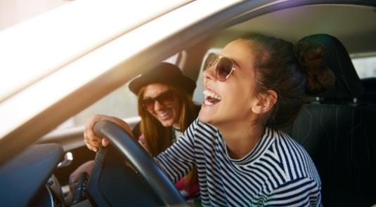 Due donne sorridenti in auto, indossano occhiali da sole, godendosi un momento felice.