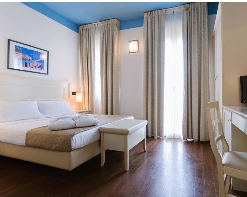 Camera d'albergo elegante, letto matrimoniale, tende lunghe e arredo chiaro.